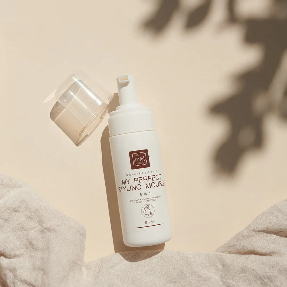 Ein weißer Flakon von Thats me Organic® Bio-Haarschaum Styling Mousse steht auf einer beigen Fläche mit sanften Schatten und einem Stoff in der Ecke, der natürliche Inhaltsstoffe für die sanfte Haarpflege präsentiert.