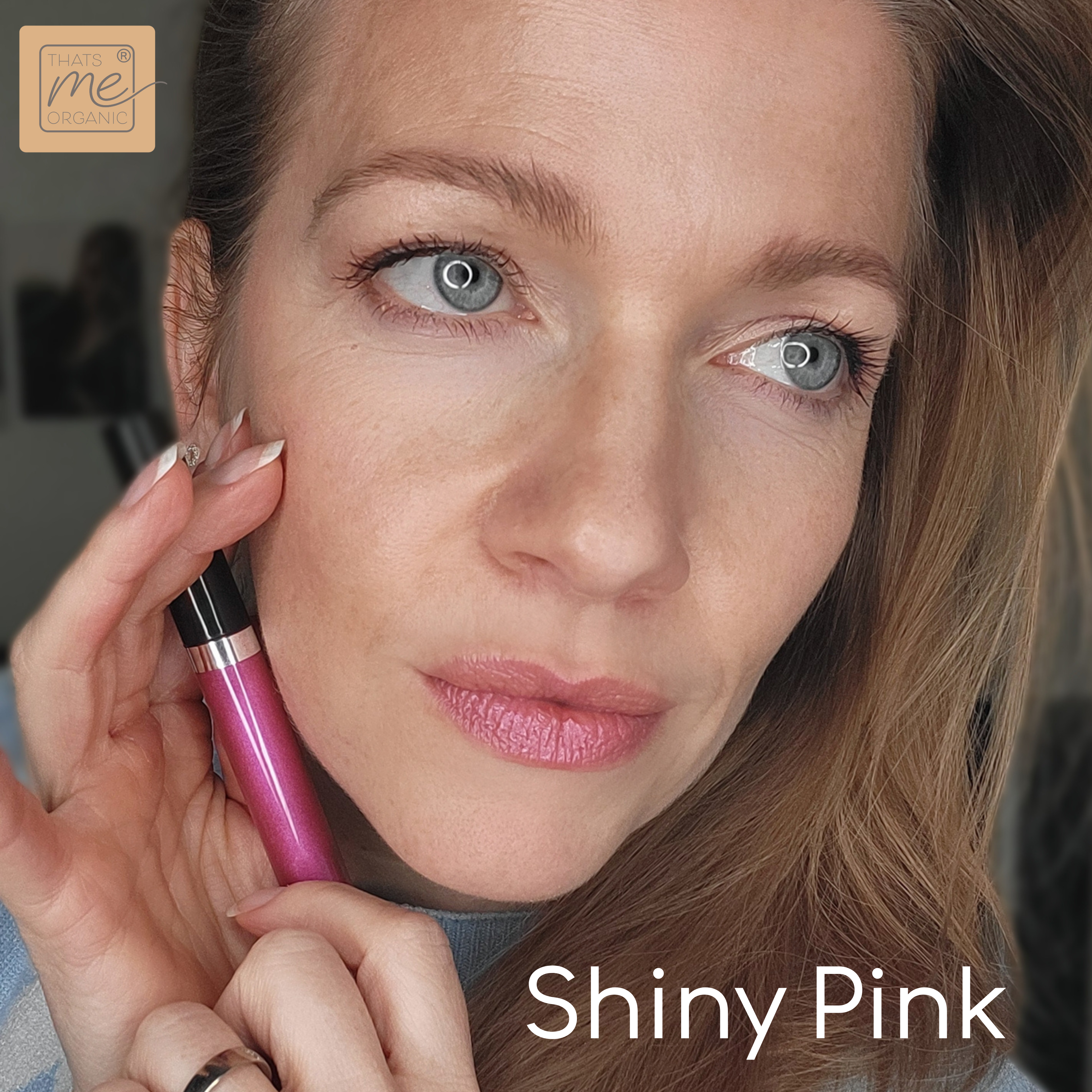 Eine Frau hält einen rosafarbenen Lippenstift in der Hand und trägt dazu passende glänzende rosafarbene Lippen, mit Thats me Organic® veganer Lipgloss für natürliche Lippenpflege. Text: Leuchtendes Pink.