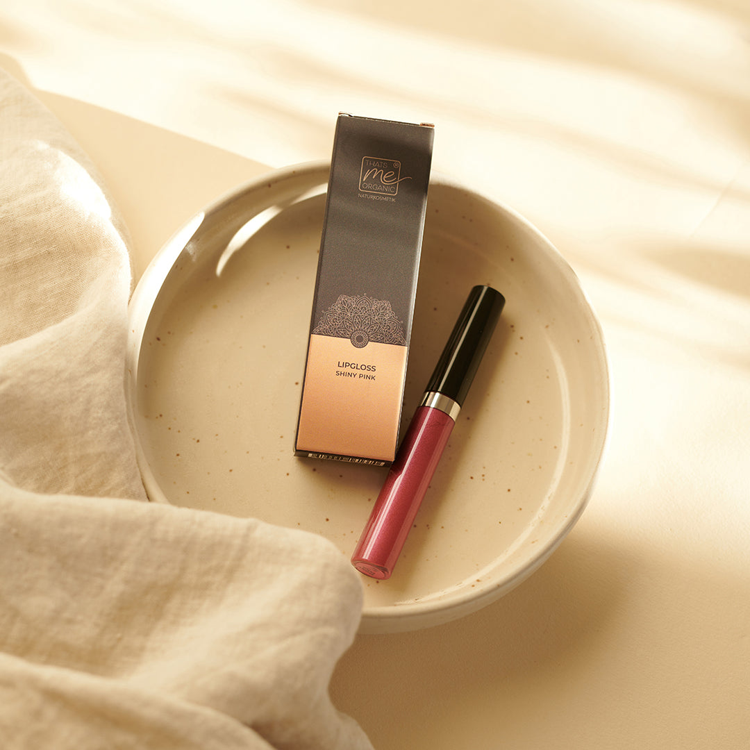 Ein rosafarbener Thats me Organic® veganer Lipgloss und seine Dose werden auf einem beigen Keramikteller neben weichem cremefarbenem Stoff präsentiert und bieten natürliche Lippenpflege bei jeder Anwendung.