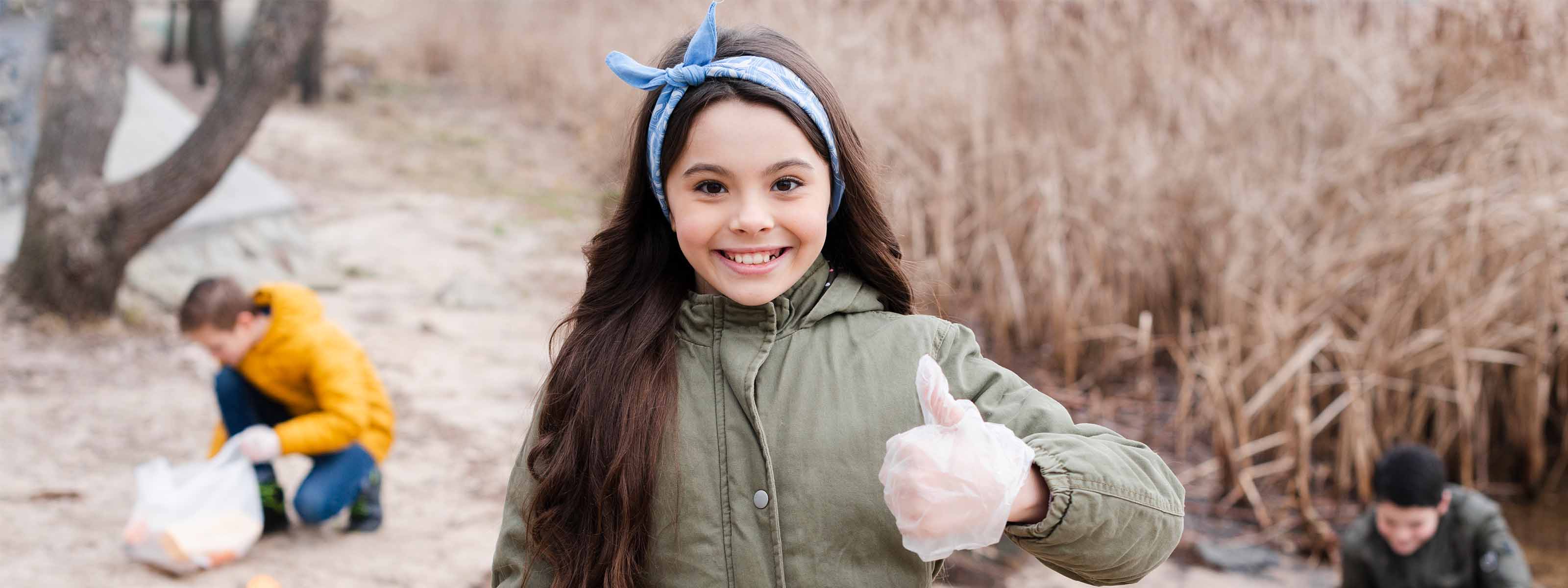 Ein lächelndes Mädchen mit Handschuhen zeigt den Daumen nach oben im Freien; Kinder räumen im Hintergrund Müll auf.