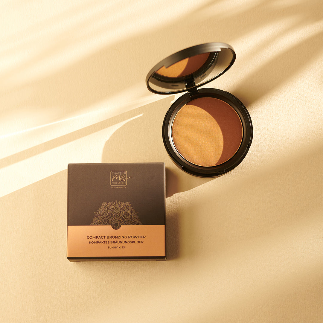 Thats me Organic® Compact Bronzing Powder mit natürlichen Inhaltsstoffen, ausgestellt in einem offenen Etui und einer Schachtel auf beigem Hintergrund, beleuchtet von sanftem Sonnenlicht.