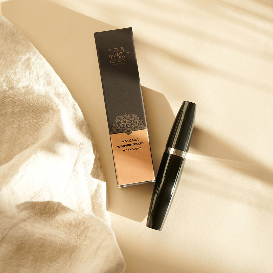 Eine schwarze Thats me Organic® Mega Volume Mascara-Tube und die dazugehörige Schachtel werden auf einem cremefarbenen Stoff in sanftem Sonnenlicht zur Schau gestellt und unterstreichen die Eleganz dieser Bio-Wimperntusche.