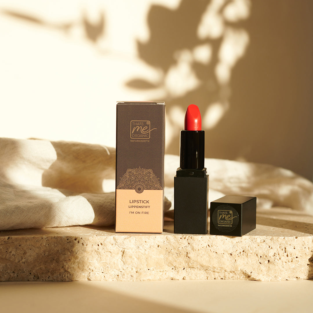 Ein roter Lippenstift von Thats me Organic® in einer schwarzen Tube mit seiner Schachtel, ausgestellt auf beigem Stein unter warmem Naturlicht - perfekt für natürliche Lippenpflege und inspiriert von tierversuchsfreien Lippenstiften.