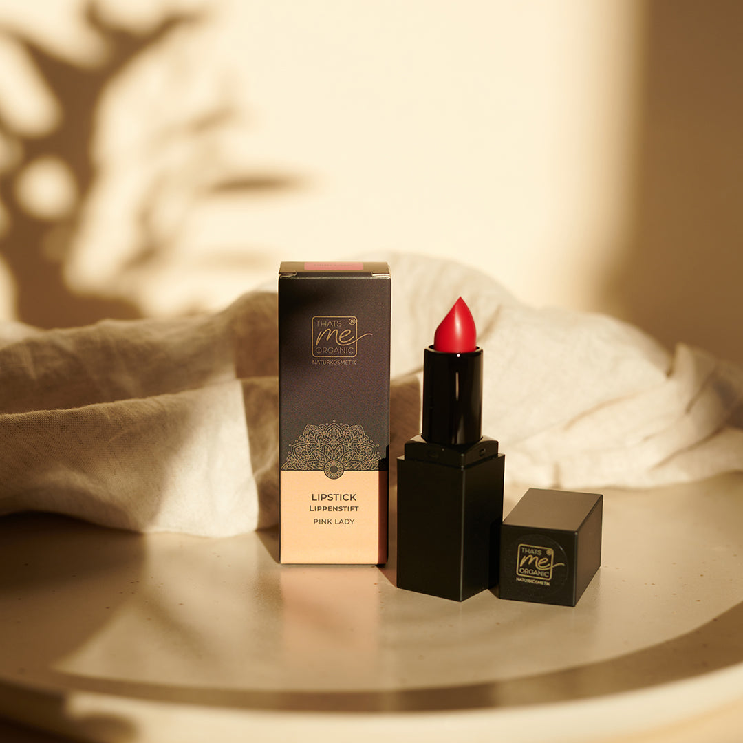 Ein roter, veganer Bio-Lippenstift von Thats me Organic® liegt neben seiner Box auf beigem Untergrund, umgeben von sanftem Sonnenlicht und zarten Schatten.