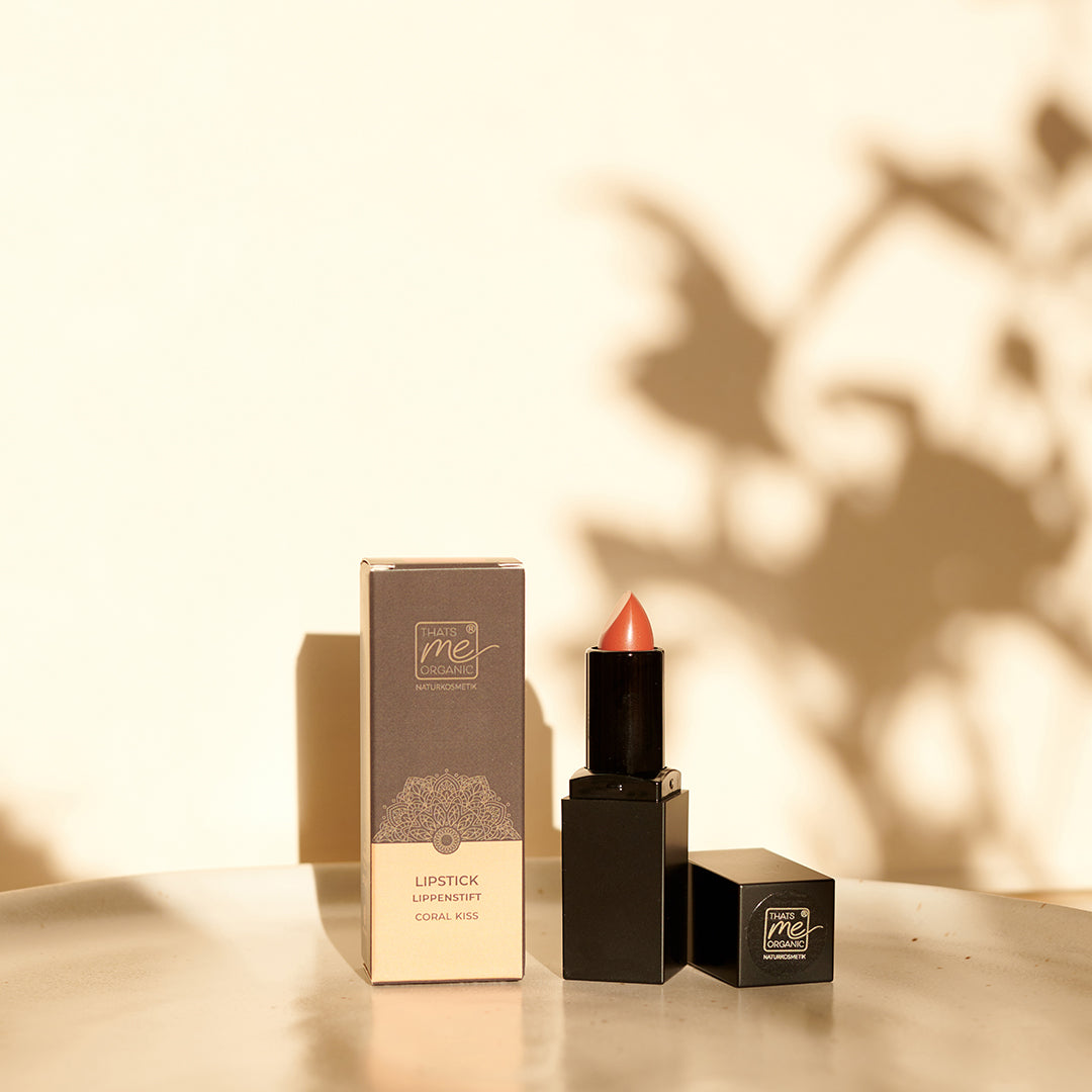 Ein korallenroter Thats me Organic®-Lippenstift und seine Verpackungsbox werden auf einem Tisch mit sanftem Sonnenlicht und Pflanzenschatten ausgestellt und unterstreichen die natürliche Lippenpflege für strahlende, gesunde Lippen.