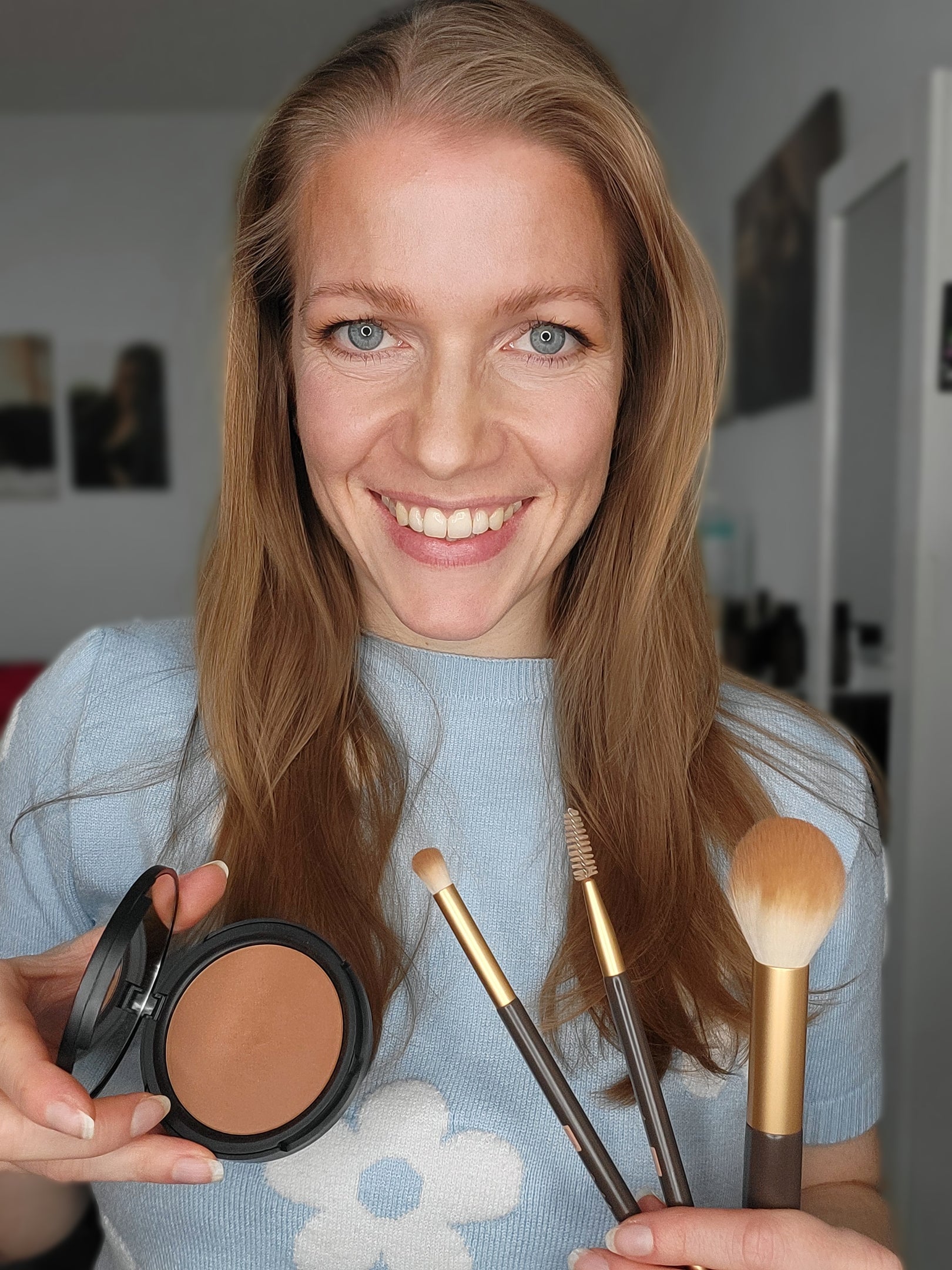Eine lächelnde Frau, die Make-up-Pinsel und Kompaktpuder in der Hand hält und ein blaues Hemd mit einer weißen Blume trägt - perfekt für die Präsentation von Thats me Organic® vegan Bronzer aus natürlichen Inhaltsstoffen.