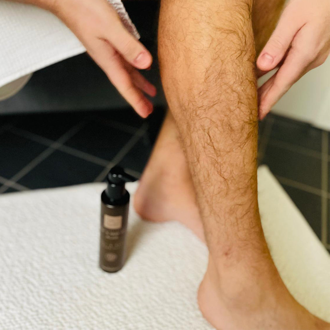 Eine Person trägt Lotion auf ihr nacktes Bein auf, während die Thats me Organic® Männerpflege Lotion und das Deo-Mousse aus einem Geschenk-Set auf einem weißen Handtuch liegen.