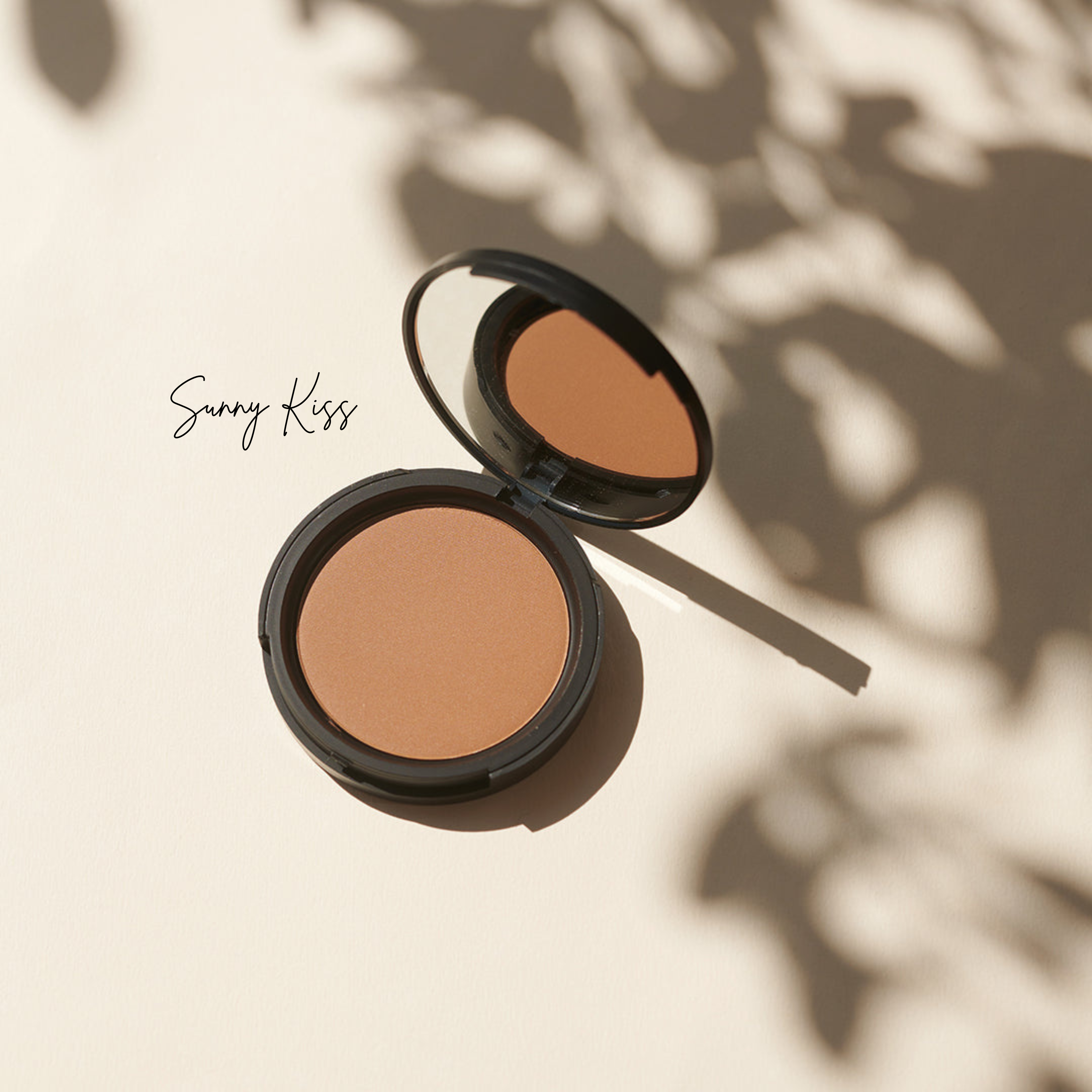 Ein offener Thats me Organic® Bronzing Powder in Sunny Kiss, hergestellt mit natürlichen Inhaltsstoffen, liegt zwischen zarten Blattschatten auf einem hellen Hintergrund.