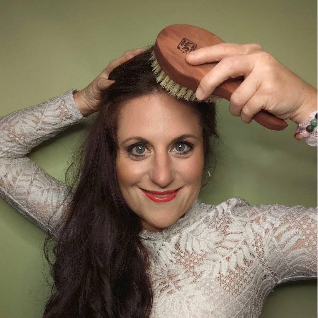 Eine Frau mit langen braunen Haaren lächelt, während sie eine Thats me organic® vegane Haarbürste auf ihrer Kopfhaut anwendet, um das Haarwachstum zu fördern, vor einem grünen Hintergrund.