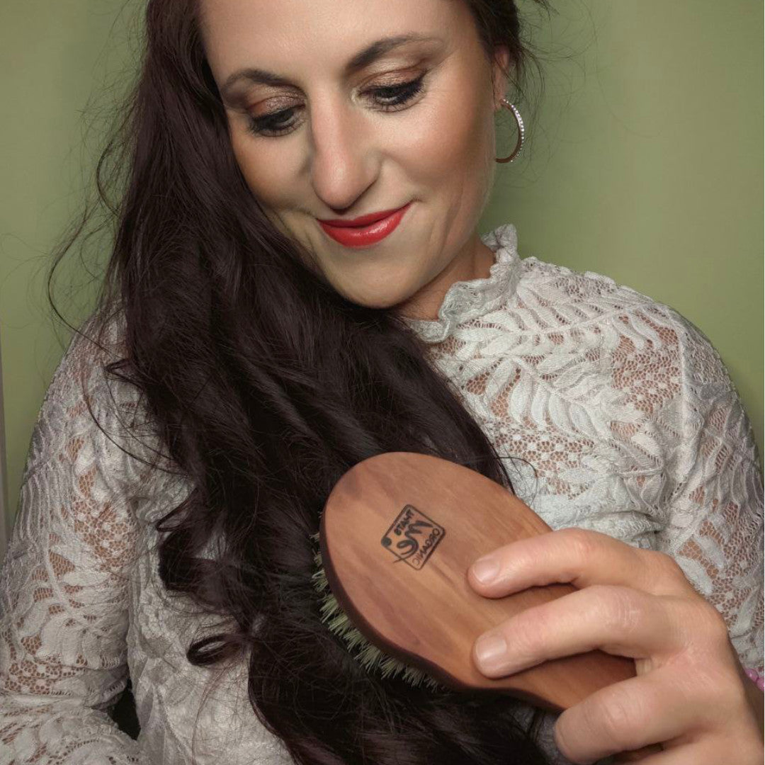 Eine Frau mit langem, dunklem Haar bürstet es lächelnd mit der Thats me organic® Naturhaarbürste Sisal, um es sanft zu entwirren und das Haarwachstum zu fördern. Sie trägt ein weißes Spitzenoberteil vor einem grünen Hintergrund.