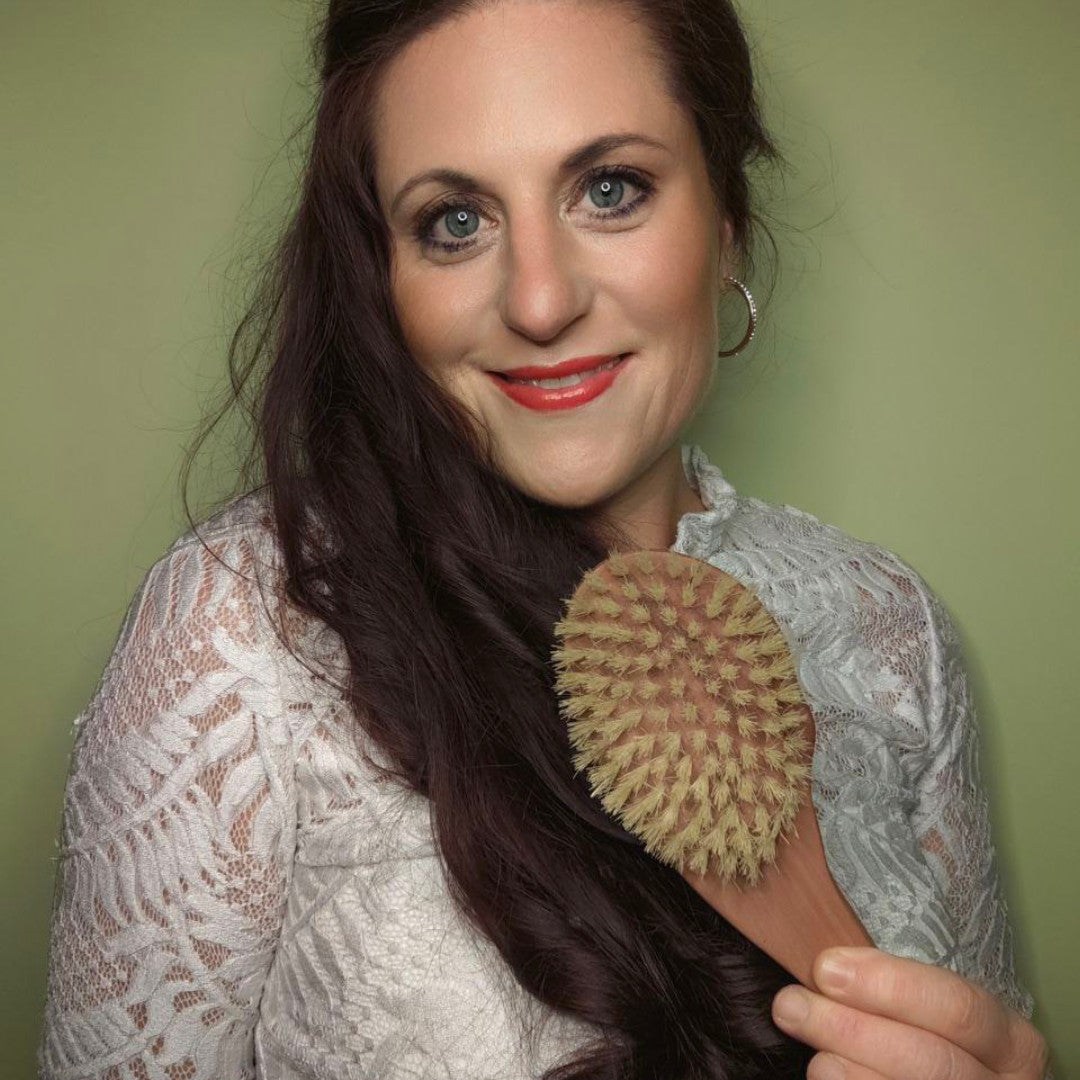 Eine Frau in einem weißen Spitzentop hält eine Thats me Organic® Tropfenform Bürste und lächelt in die Kamera vor einem grünen Hintergrund. Diese einzigartig geformte Bürste unterstützt das Haarwachstum und ist somit ideal für Ihre Haarpflege.