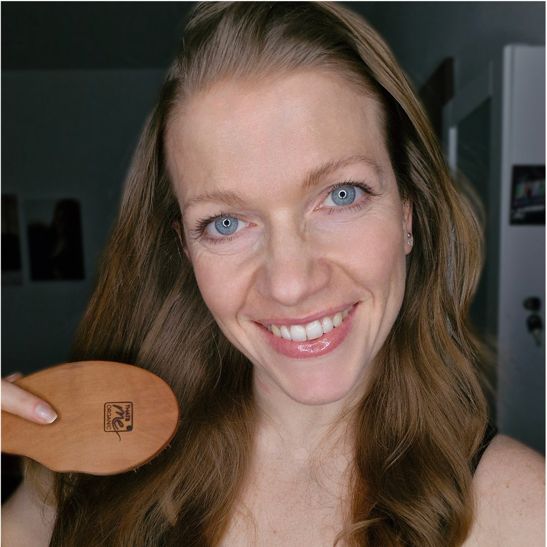 Eine lächelnde Frau mit langen braunen Haaren steht im Haus und hält eine Thats me organic® vegane Haarbürste in der Hand, bereit, Haarwachstum zu fördern.
