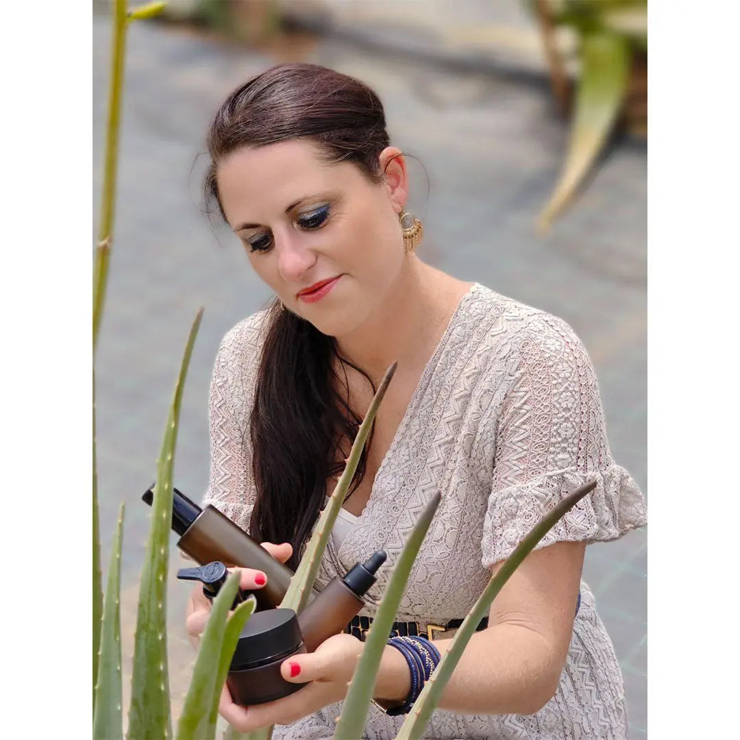 Eine Frau in einem hellen Kleid untersucht eine braune Flasche Thats me organic® Bio-Hand & Body Wash, während sie zwischen Aloe-Vera-Pflanzen sitzt.