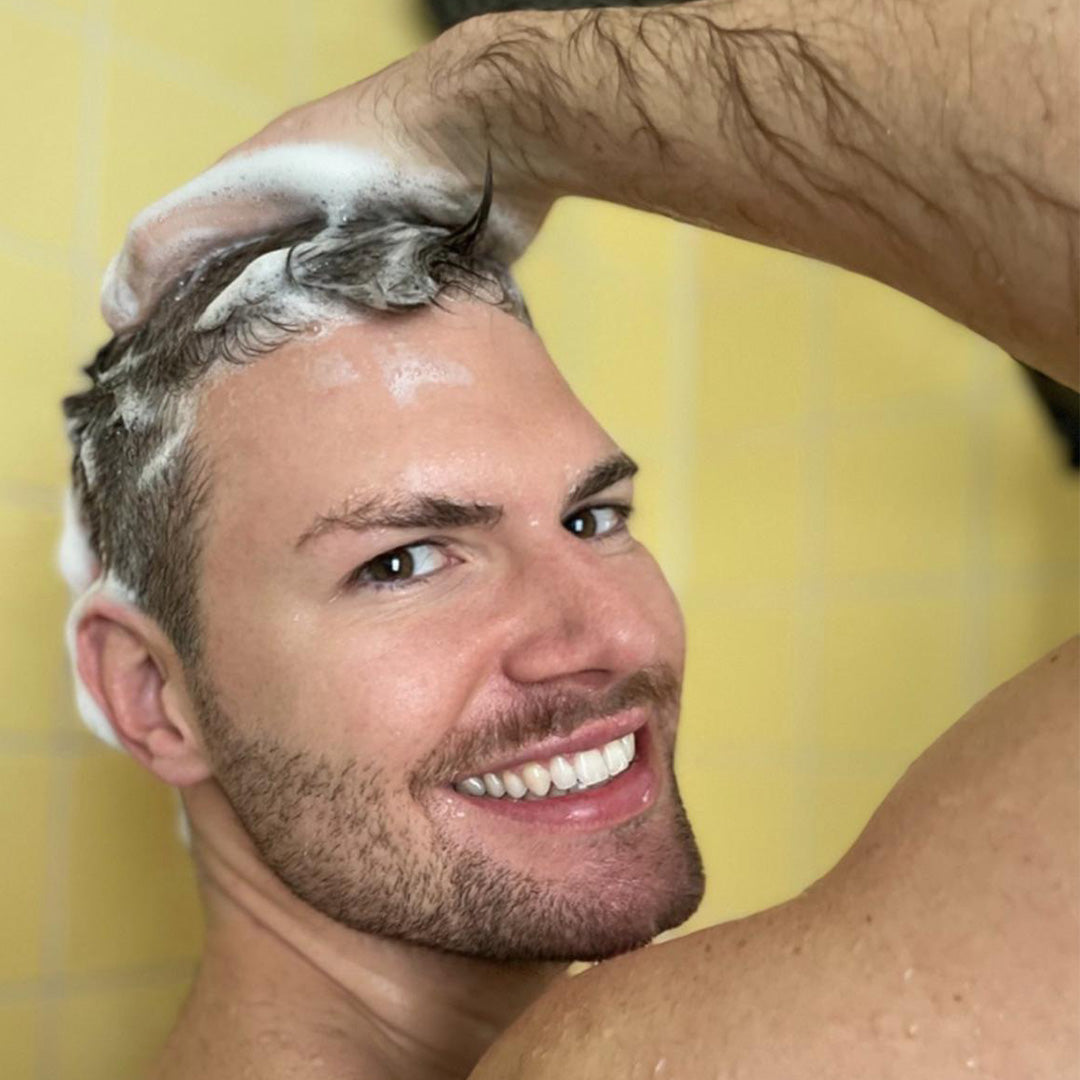Ein lächelnder Mann wäscht sich die Haare mit Thats me Organic® Shampoo unter der Dusche, schaut über die Schulter in die Kamera und genießt die natürliche Männerpflege als Teil seiner täglichen Routine.