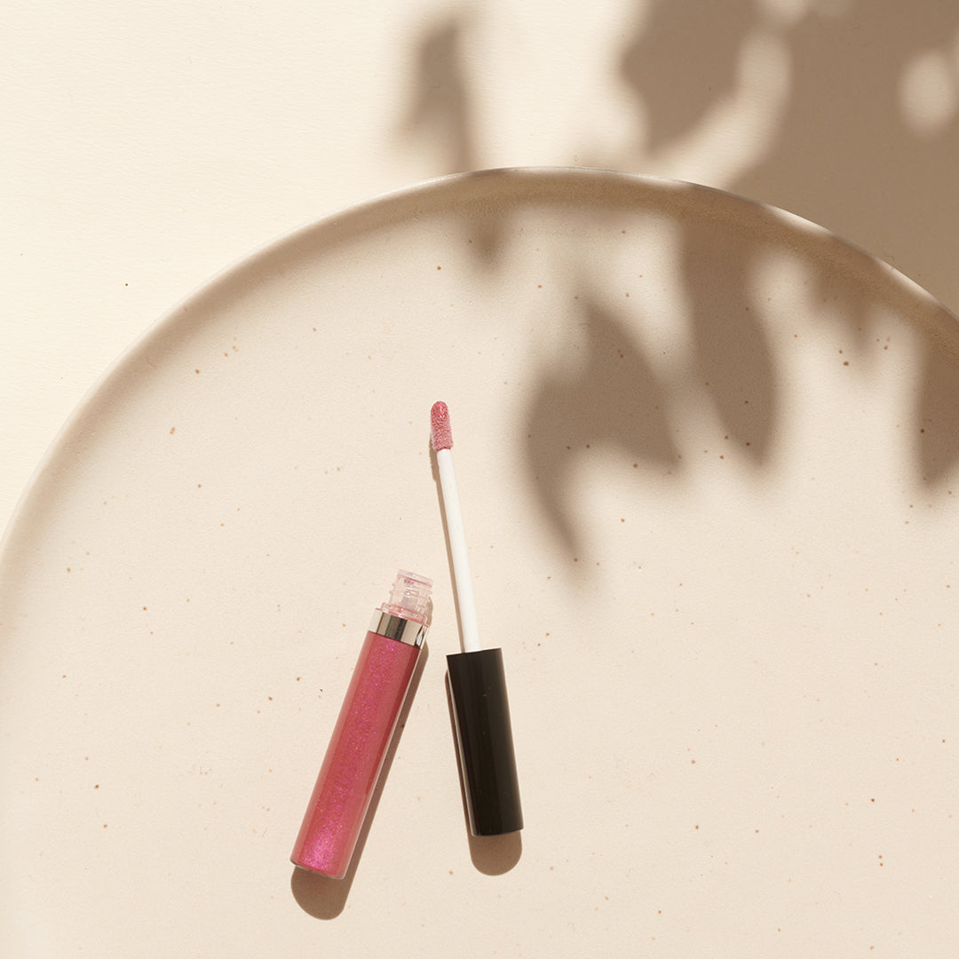Veganer Lipgloss von Thats me Organic® mit schwarzem Deckel und Applikator auf beigem Teller, umgeben von sanften Schatten - für natürliche Lippenpflege mit stilvollem Glanz.