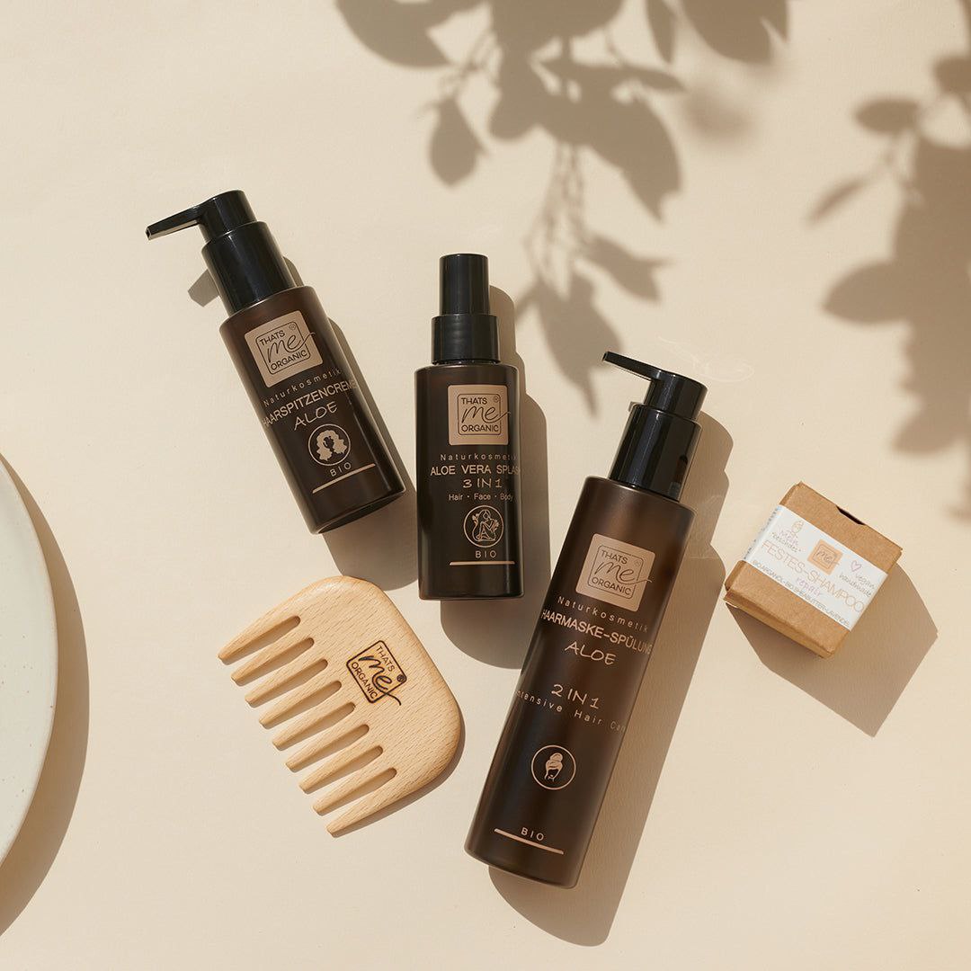 Natürliche Haarpflegeprodukte und ein hölzerner Kamm, ordentlich auf einer hellbeigen Fläche mit weichen Schatten angeordnet.