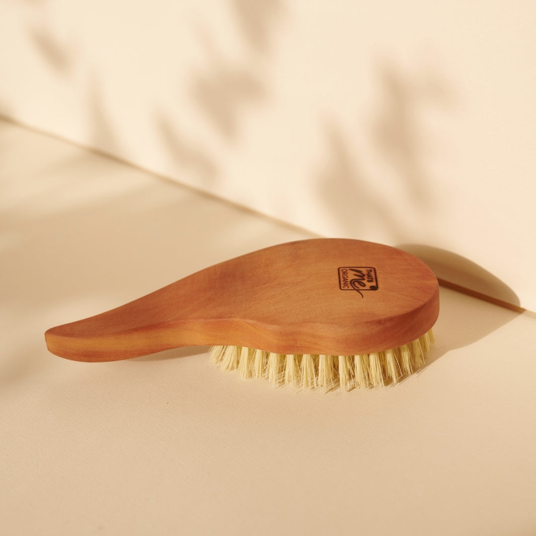 Eine vegane Thats me Organic® Haarbürste in Tropfenform mit hellen Borsten liegt auf einer beigen Oberfläche im sanften Sonnenlicht.