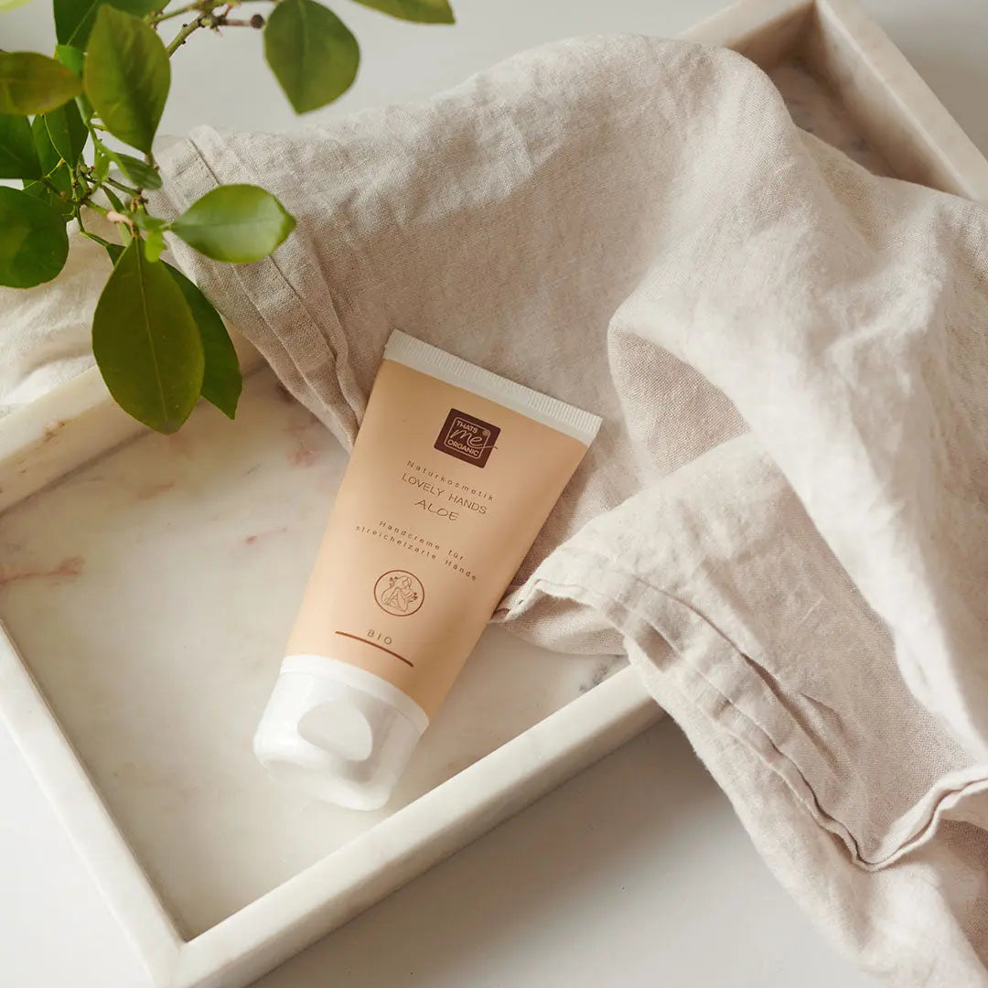 Eine Tube Thats me Organic® Bio-Naturkosmetik Handcreme mit Hyaluron steht auf einem Marmortablett, dazu ein beiges Tuch und frische grüne Pflanzenblätter.