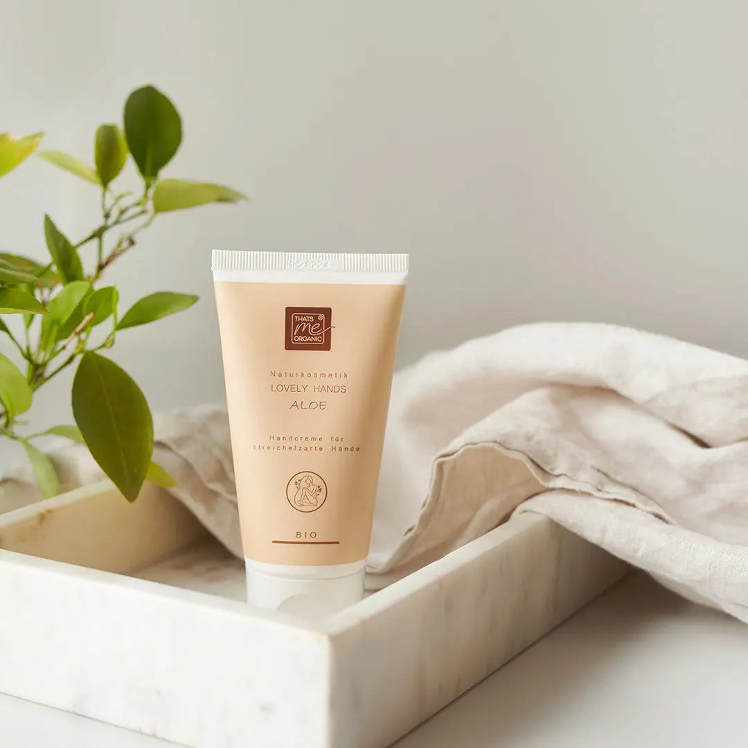 Eine Tube Thats me Organic® Hyaluron Handcreme liegt in einem Marmortablett neben einer grünen Pflanze und einem beigen Tuch.