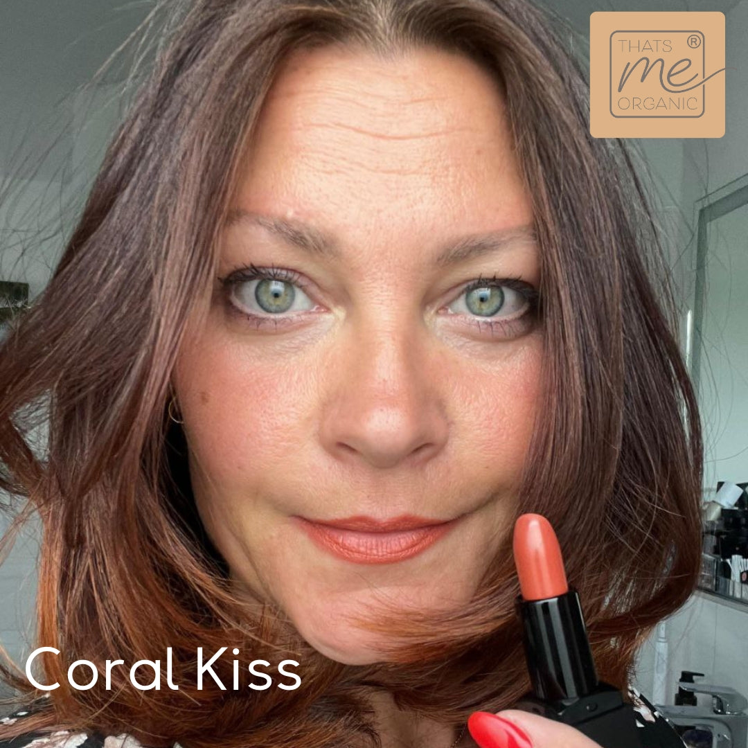 Eine grünäugige Frau trägt korallenroten Lippenstift und hält dazu passende tierversuchsfreie Lippenstifte: Coral Kiss von Thats me Organic®.