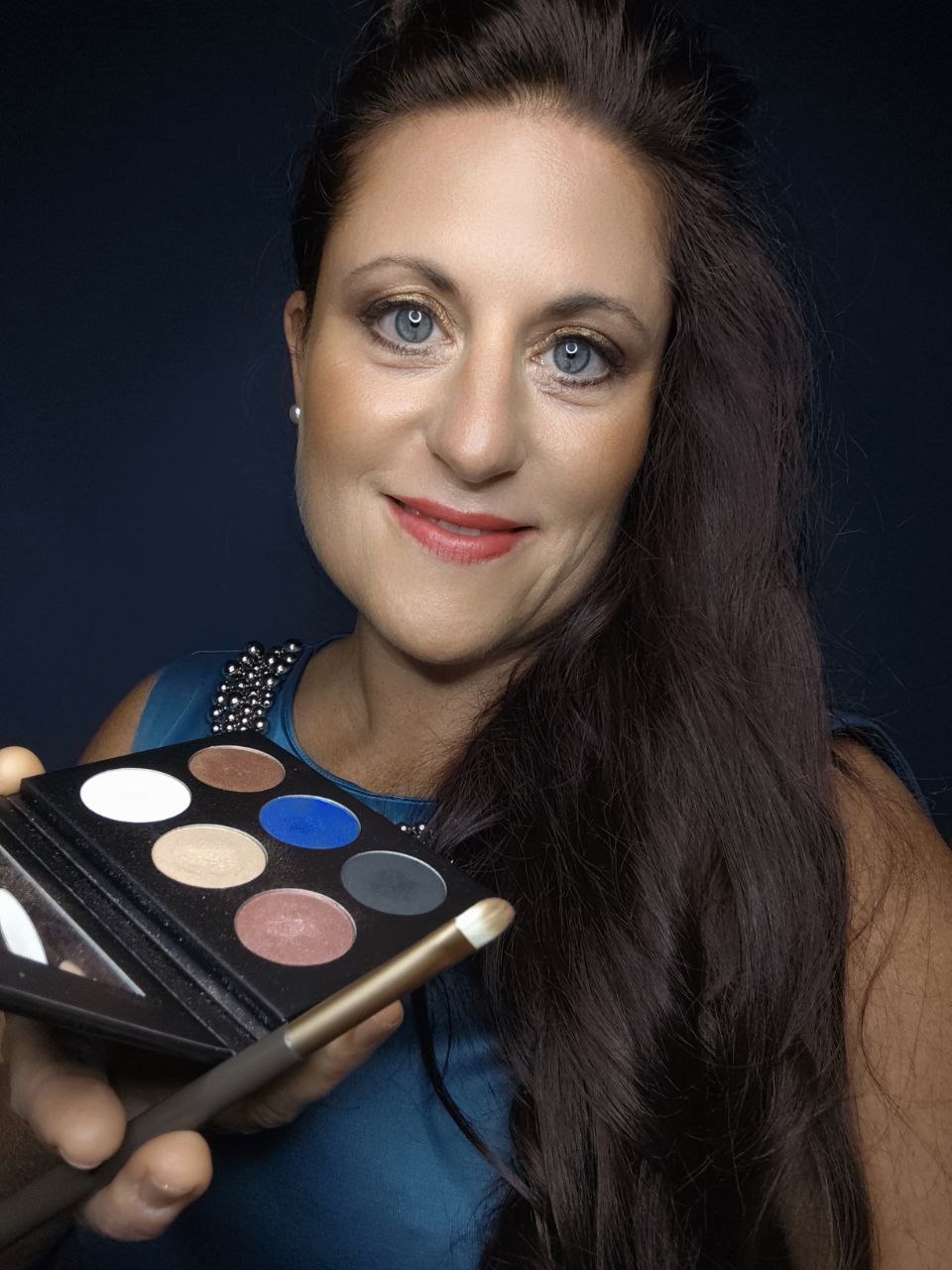 Eine Frau mit langen dunklen Haaren lächelt in die Kamera und hält eine Thats me Organic® 6 in 1 Lidschattenpalette und einen Pinsel vor einem dunklen Hintergrund.