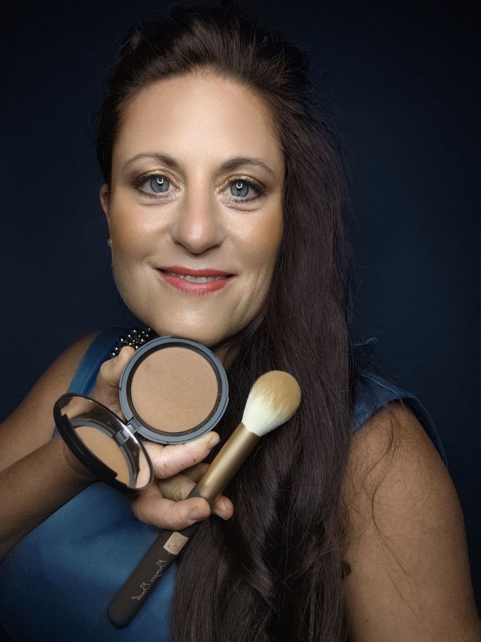 Eine lächelnde Frau hält eine Thats me Organic® Bronzing Powder Compact und einen Make-up-Pinsel vor einem dunklen Hintergrund, der den mühelosen Glanz dieses veganen Bronzers unterstreicht.