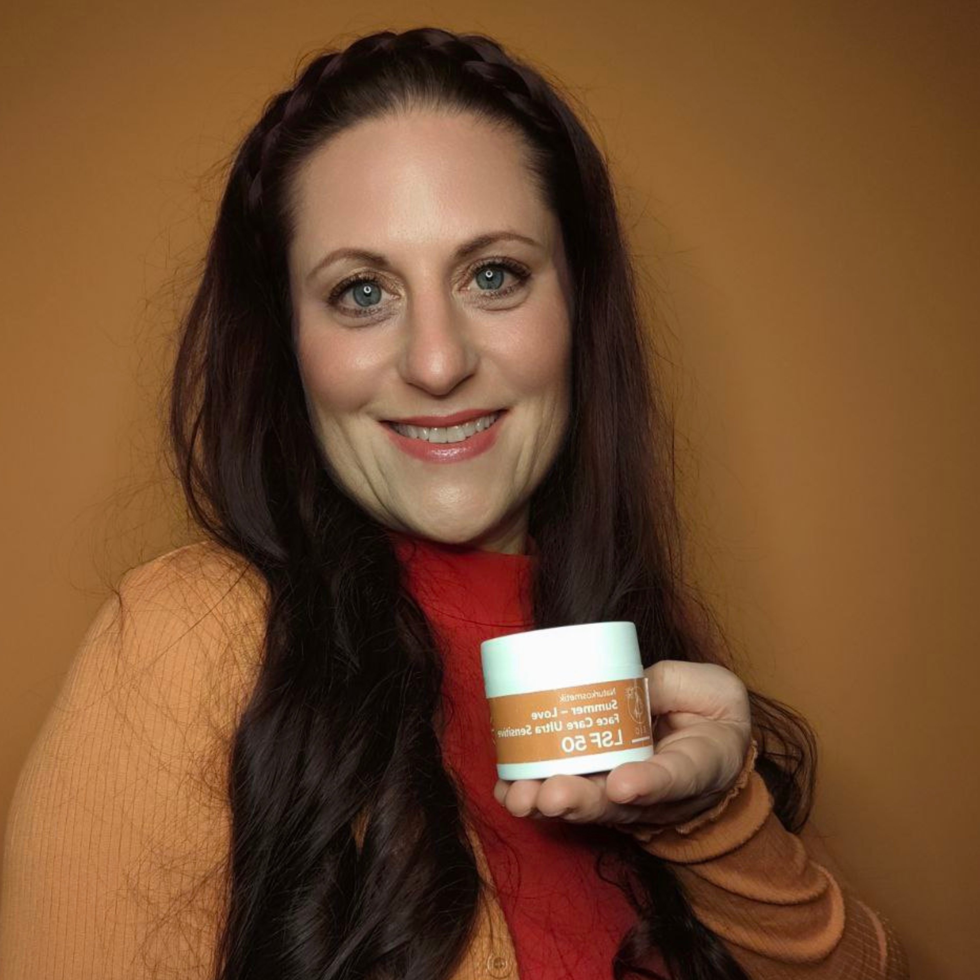 Lächelnde Frau mit langen dunklen Haaren hält einen Thats me Organic® Sonnencreme LSF 50+ Tiegel, der UVA und UVB Schutz hervorhebt, vor einem orangen Hintergrund.