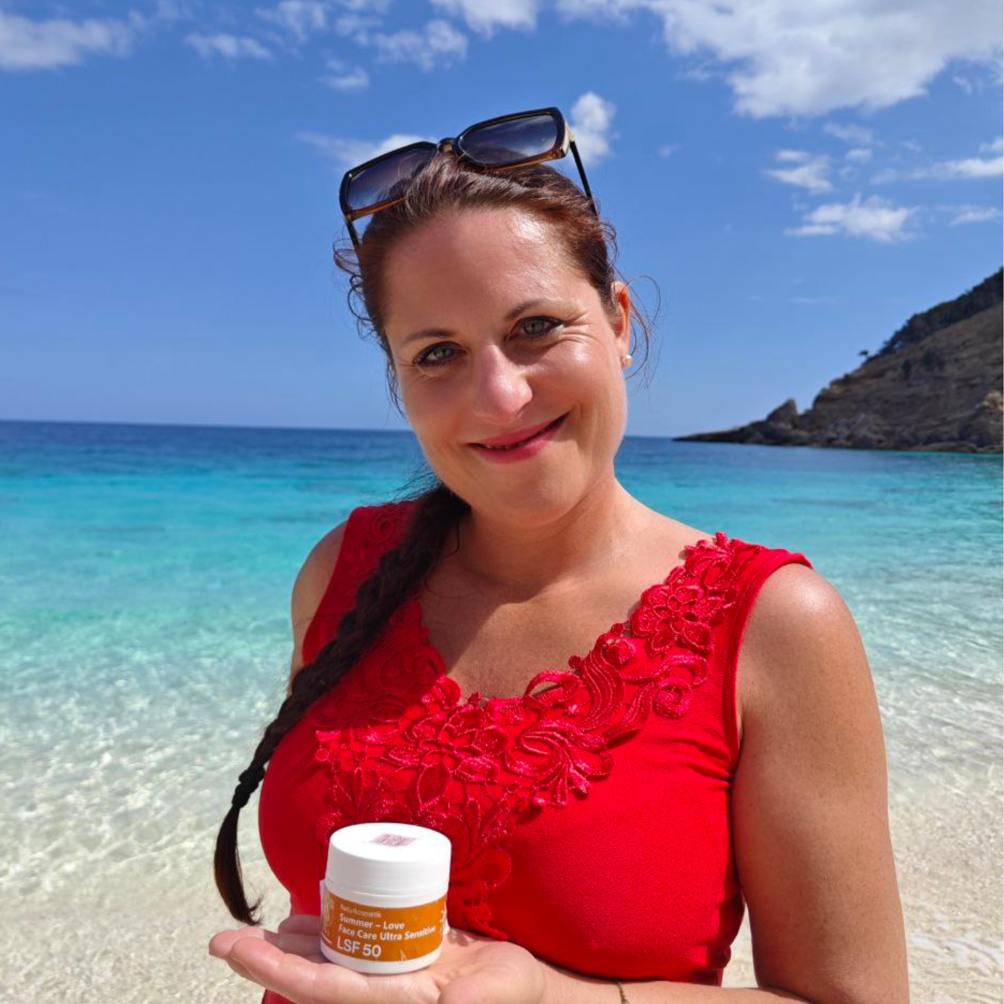 Frau in rotem Top mit Thats me Organic® Sonnencreme LSF 50+ ohne Titandioxid an einem sonnigen Strand, mit blauem Meer und Himmel im Hintergrund.