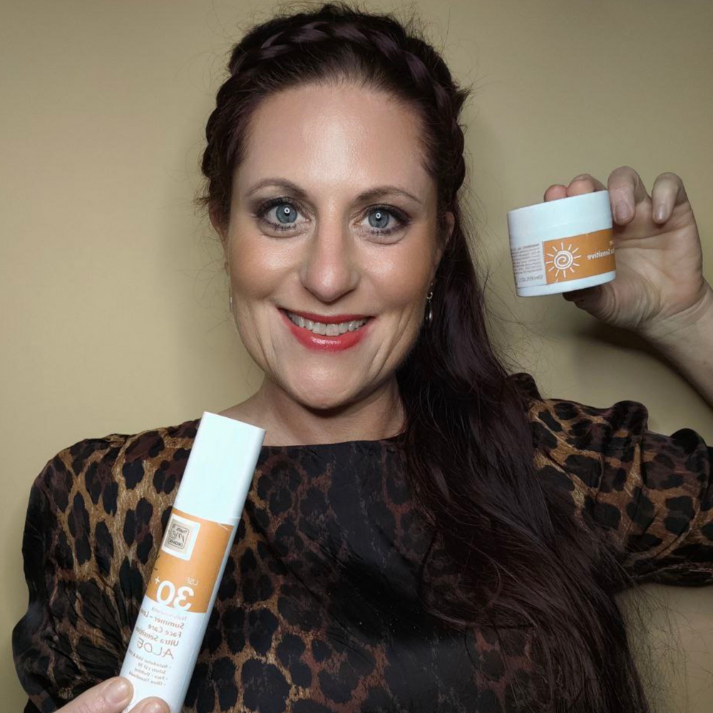 Eine lächelnde Frau mit Leopardenmuster hält eine Thats me Organic® Bio Sonnencreme LSF 30+ Flasche und einen Hautpflegetiegel mit der Aufschrift Sonnenschutz ohne Titandioxid vor einem beigen Hintergrund.