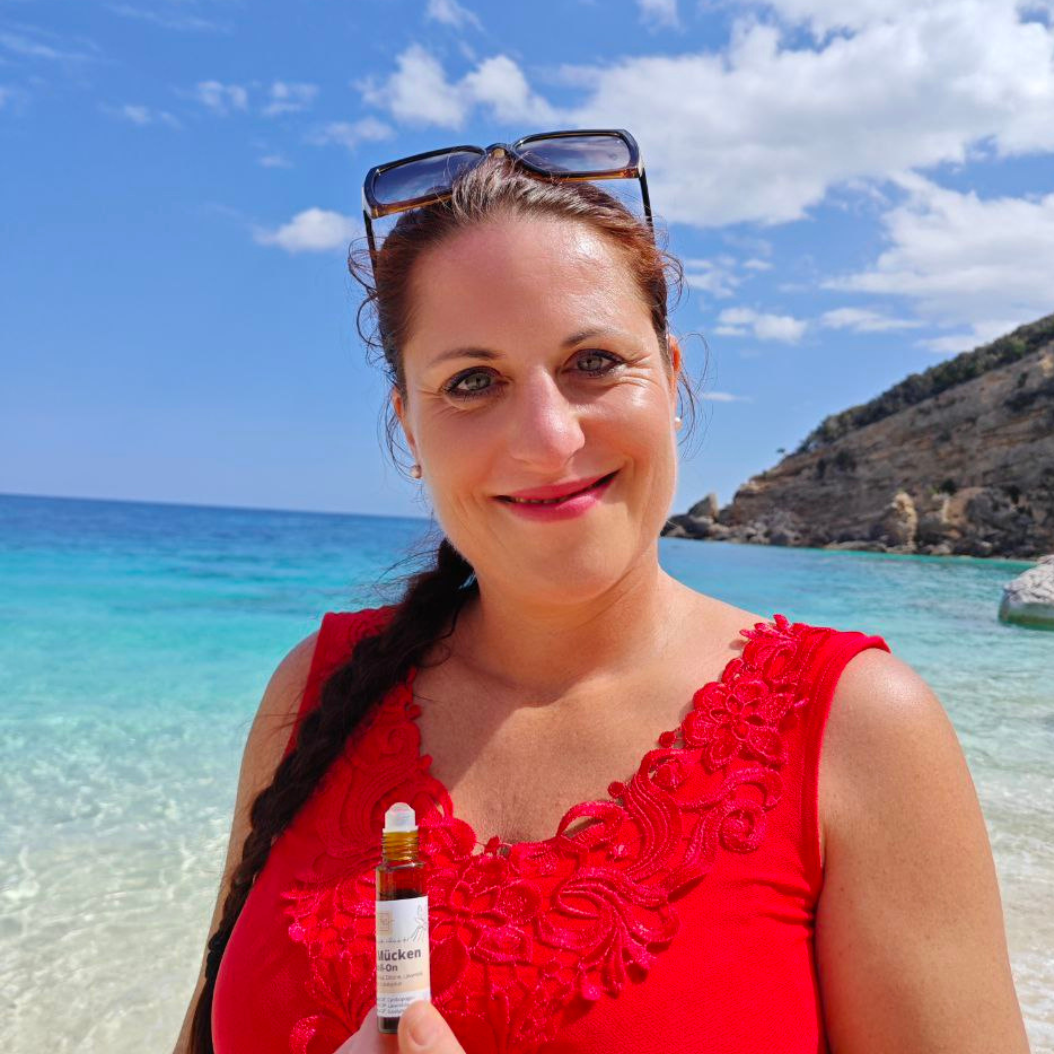 Eine Frau in einem roten Kleid lächelt am Strand, in der Hand eine kleine Tube Thats me Organic® Anti-Mücken-Roll-On, mit blauem Meer und Himmel im Hintergrund - ein perfekter Moment des natürlichen Mückenschutzes.