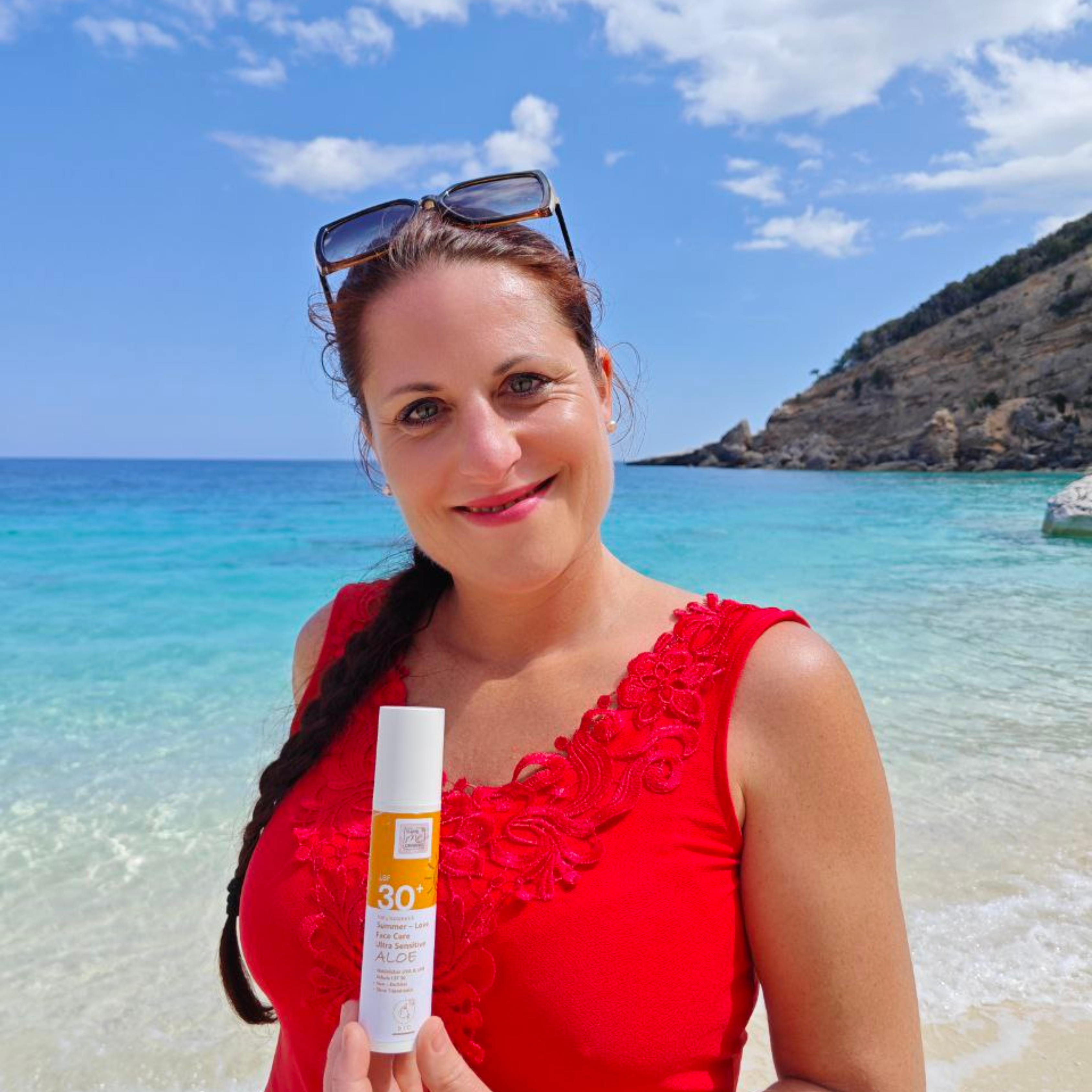 Eine Frau im roten Kleid hält Thats me Organic® Bio Sonnencreme LSF 30+ an einem sonnigen Strand mit klarem, blauem Wasser und genießt den effektiven Sonnenschutz ohne Titandioxid.