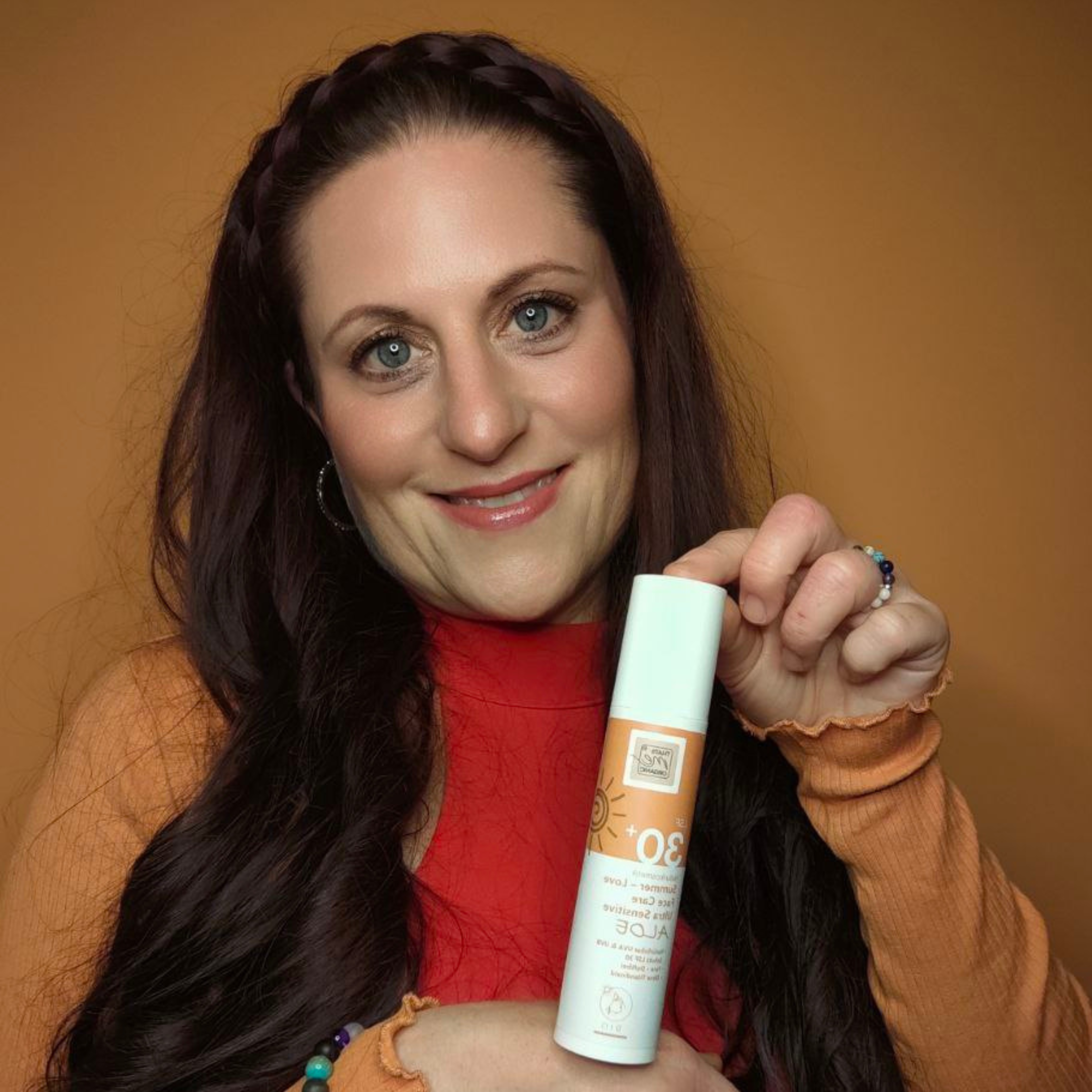 Eine Frau mit langen braunen Haaren hält eine Flasche Thats me Organic® Bio Sonnencreme LSF 30+ und lächelt vor einem orangefarbenen Hintergrund.