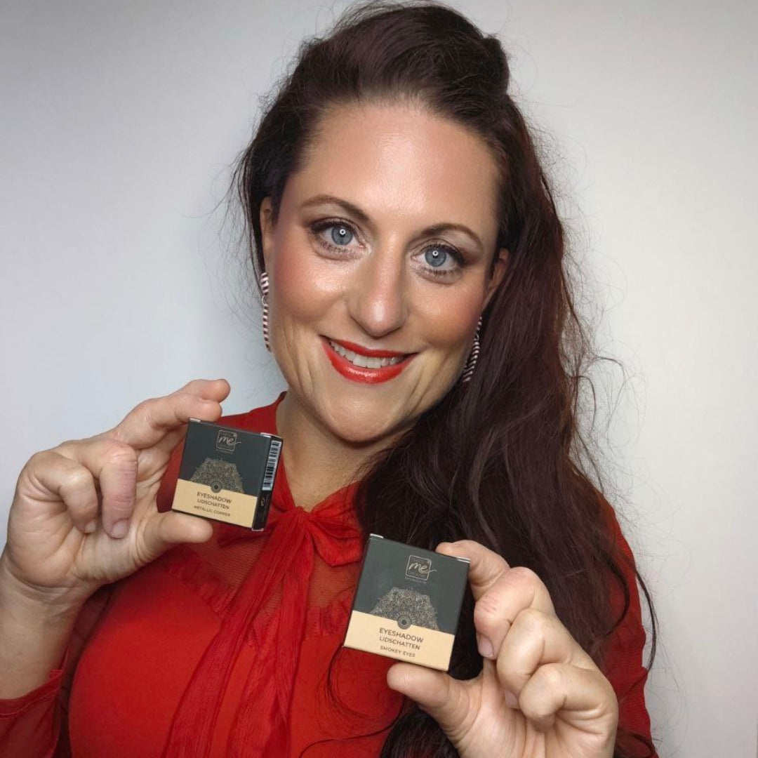 Eine Frau mit rotem Oberteil hält lächelnd zwei kleine schwarz-goldene Thats me Organic® Naturkosmetik Lidschatten-Dosen - vegan und titandioxidfrei - vor einem schlichten Hintergrund.