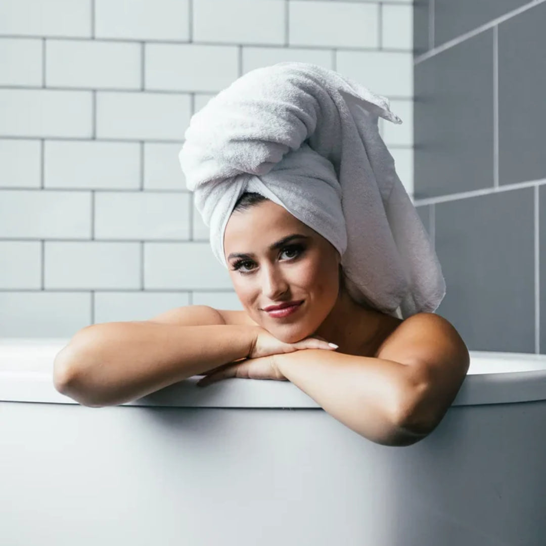 Eine Frau entspannt sich in der Badewanne, lächelnd, mit einem Handtuch um den Kopf gewickelt, und genießt den erfrischenden Duft von Thats me Organic® umweltfreundliches Shampoo.