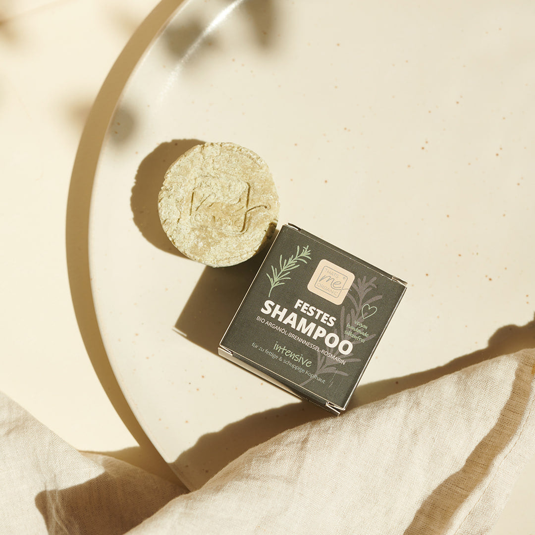 Ein runder Thats me Organic® Festes Shampoo Riegel und seine Schachtel mit der Aufschrift "Festes Shampoo" liegen auf einem beigen Teller im Sonnenlicht - ein umweltfreundliches Produkt, ideal für nachhaltige Haarpflege.