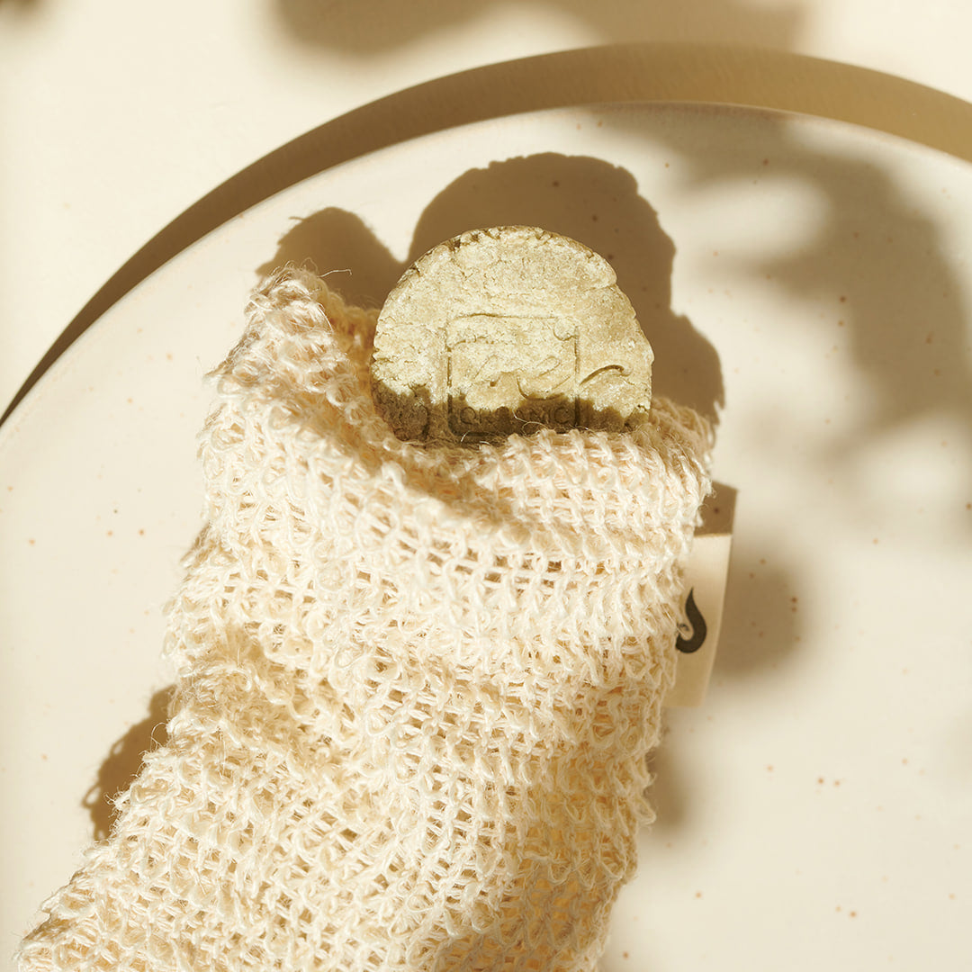 Ein runder, grüner Brennessel Rosmarin Shampoo-Riegel von Thats me Organic® liegt teilweise eingewickelt in ein strukturiertes, cremefarbenes Tuch auf einem gesprenkelten Teller - ein umweltfreundliches Shampoo für eine erfrischende, nachhaltige Haarpflege.