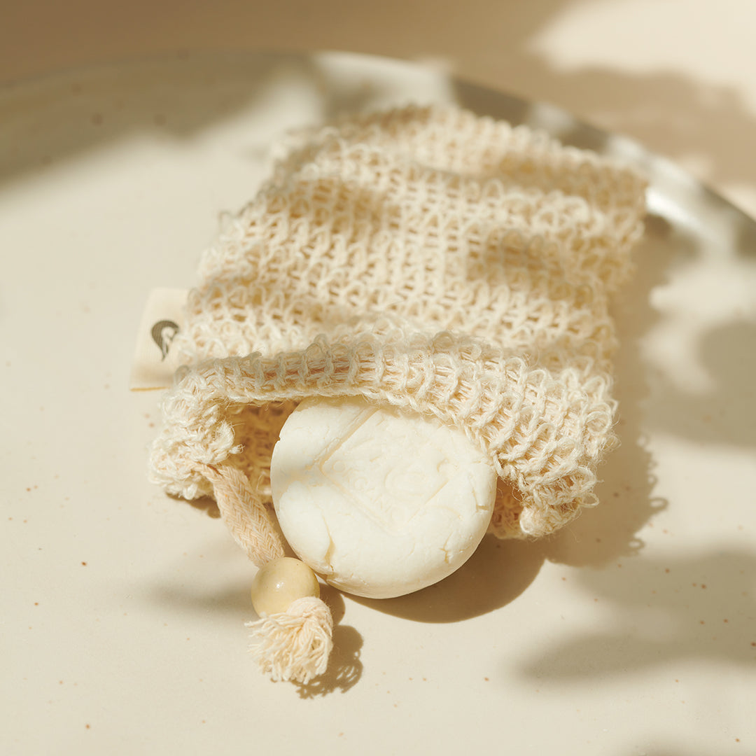 Ein weißes Thats me Organic® Shampoo Bar ruht in einem cremefarbenen, gewebten Seifensparbeutel mit einer Holzperle, die auf einer hellen Oberfläche präsentiert wird. Jetzt angereichert mit pflegendem BIO Arganöl für eine sanfte Reinigung.