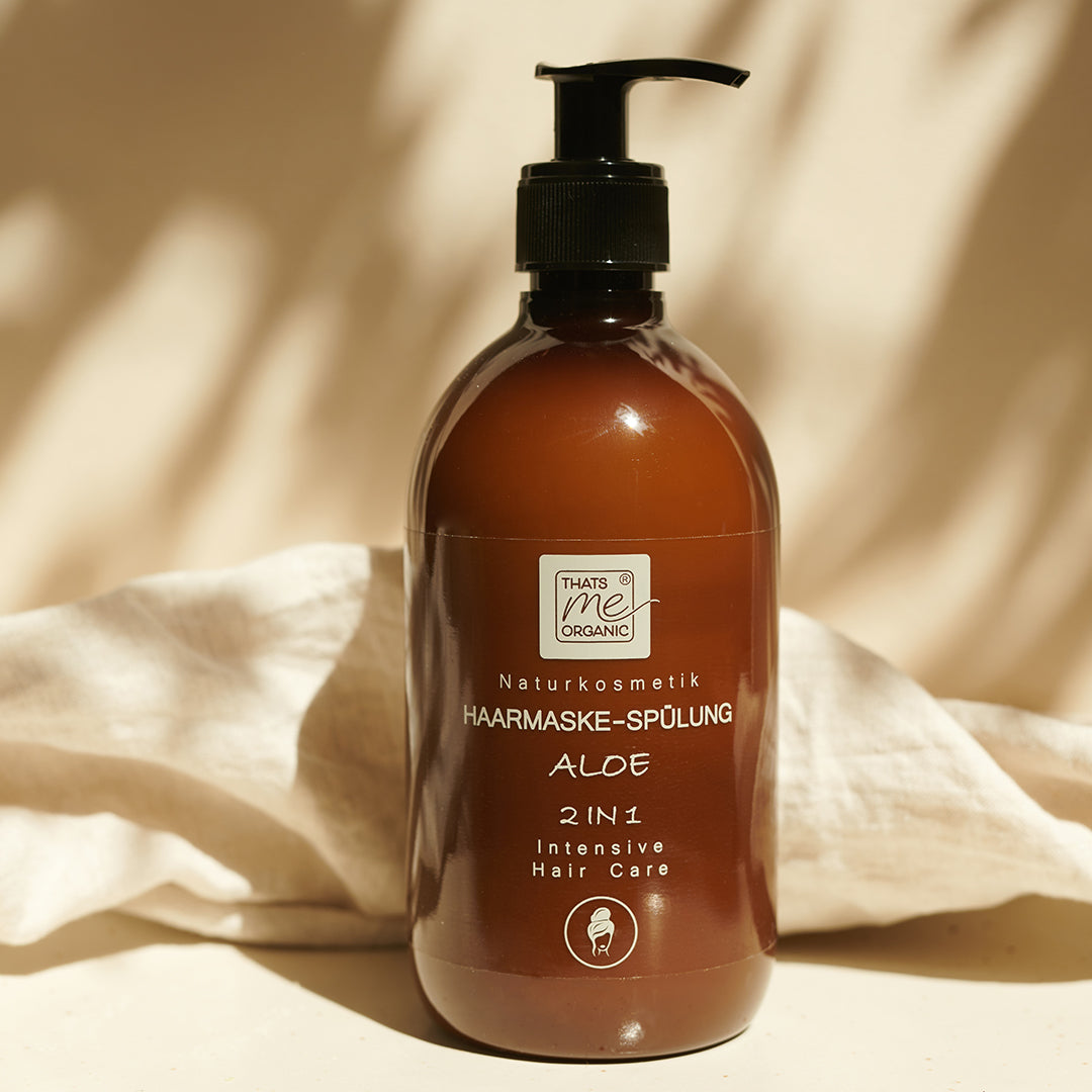 Eine braune Pumpflasche mit Thats me organic® aloe 2-in-1 intensive Bio-Haarmaske und Spülung für natürliche Haarpflege steht auf einem beigen Stoffhintergrund.