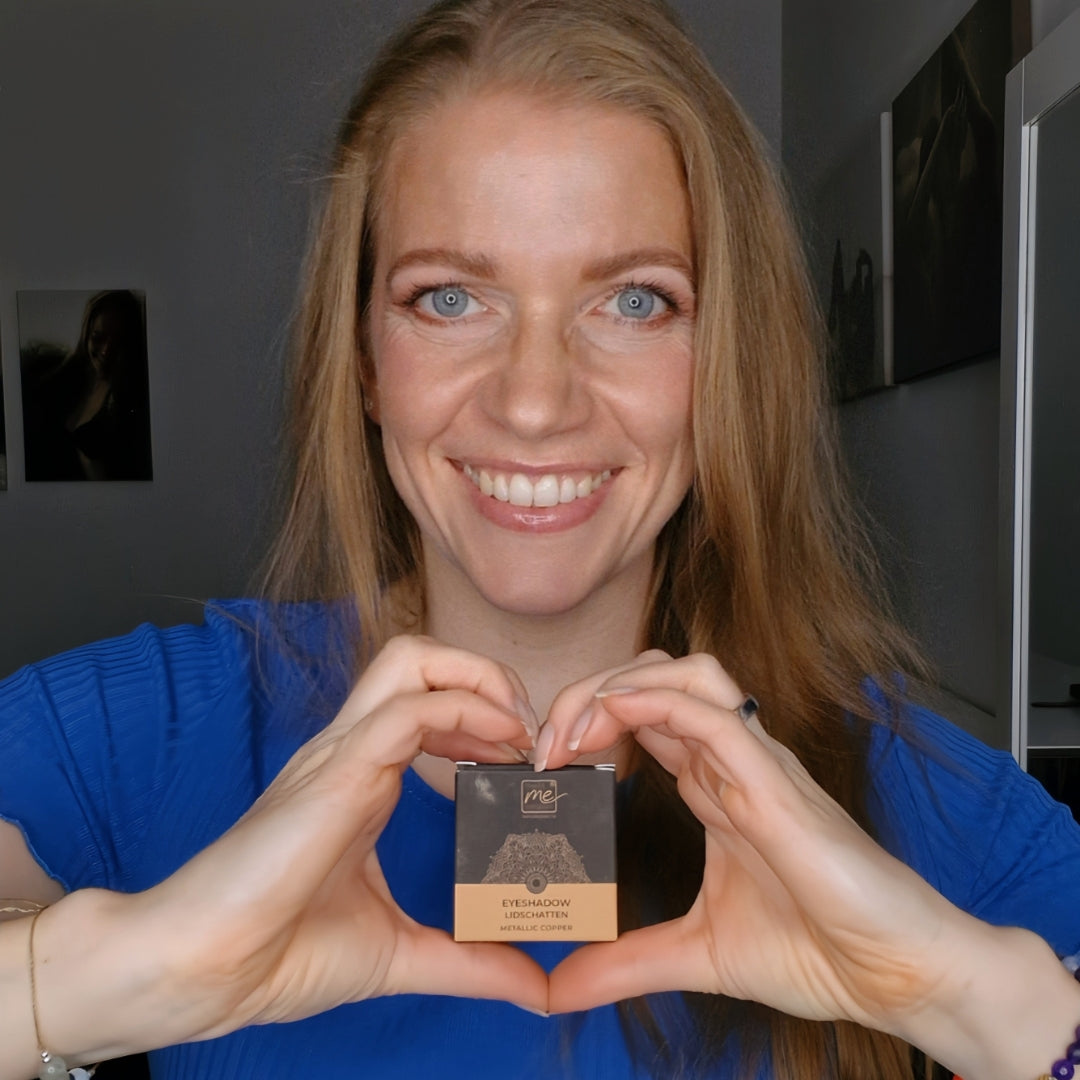 Lächelnde Frau in blauem Oberteil hält Thats me Organic® Lidschatten in Metallic Copper und formt mit ihren Händen eine Herzform um die titandioxidfreie Naturkosmetikdose.