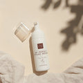 Ein weißer Flakon von Thats me Organic® Bio-Haarschaum Styling Mousse steht auf einer beigen Fläche mit sanften Schatten und einem Stoff in der Ecke, der natürliche Inhaltsstoffe für die sanfte Haarpflege präsentiert.