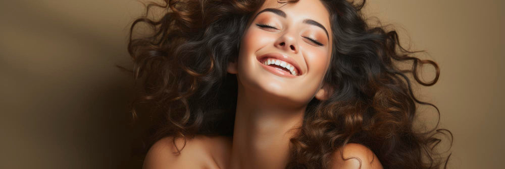 Frau mit langem, lockigem Haar, die vor einem warmen, beigen Hintergrund fröhlich lächelt.