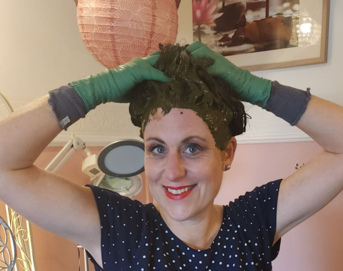 Eine lächelnde Frau mit grünen Handschuhen trägt in einem gemütlichen Raum grünes Haarfärbemittel oder grüne Paste auf ihr Haar auf.