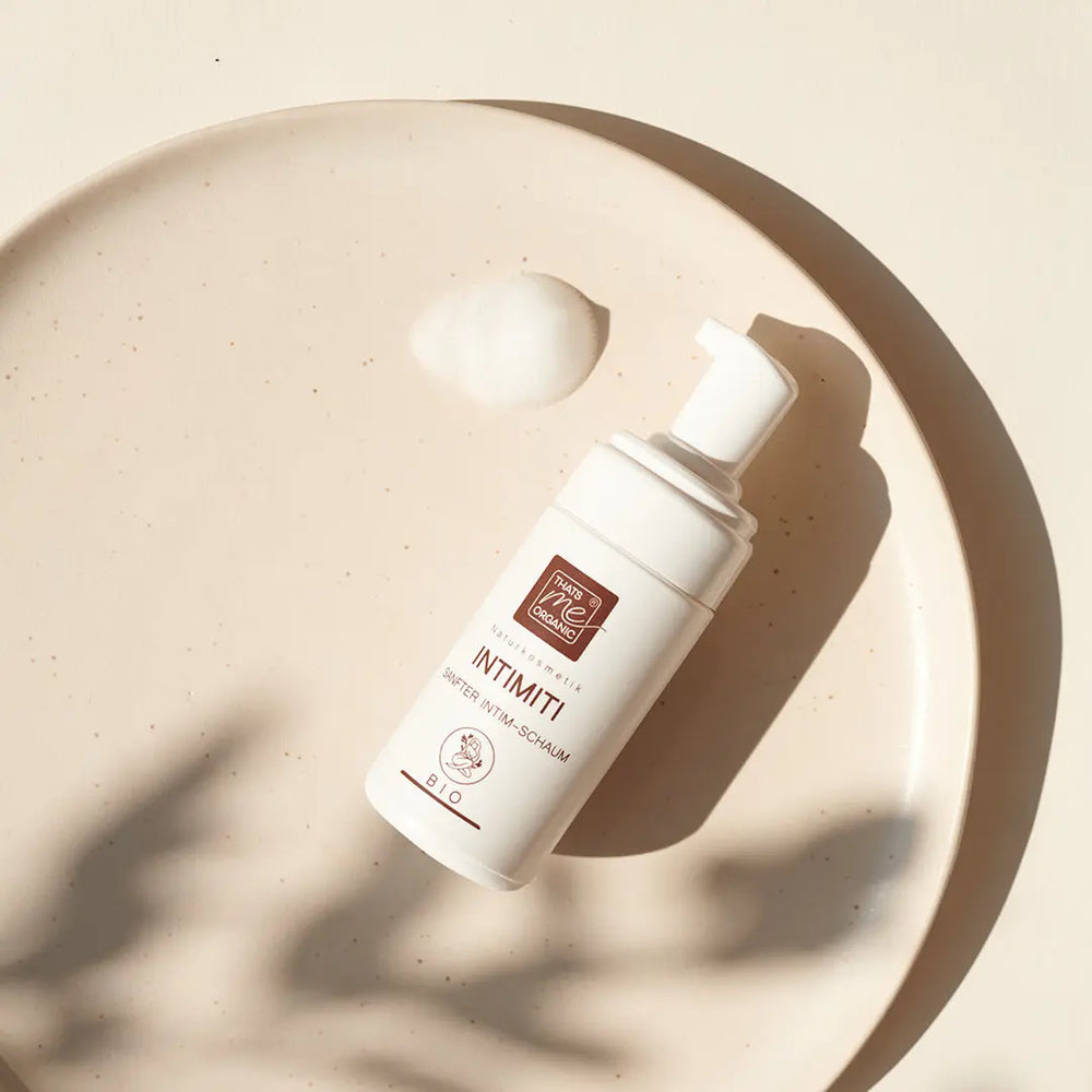Das ist die weiße Organic®-Flasche des natürlichen Intimreinigers auf einer beigen Platte mit einer Schaumwolke daneben, sanft beleuchtet vom Sonnenlicht.