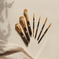 Acht Make-up-Pinsel aus dem Thats me Organic® Profi-Pinsel Set, mit goldenen und schwarzen Stielen, arrangiert auf einer hellbeigen Oberfläche mit weichen Schattierungen.