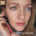 Frau mit hellen Augen, die eine rosafarbene Lipgloss-Tube in der Hand hält; in der unteren rechten Ecke steht Shiny Nude.