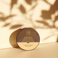 Die runde, kompakte Bio-Puderdose von Thats me Organic® mit goldenem Design wirft einen Schatten auf Beige, akzentuiert durch pflanzliche Schattierungen. Bietet einen mattierenden Effekt für einen makellosen Look.