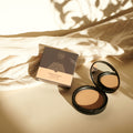 Thats me Organic® mattierendes Bio-Puder in einem eleganten, schwarz verspiegelten Etui, das neben der Schachtel auf beigem Stoff mit weichen Schatten abgebildet ist.