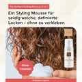 Eine Hand hält Thats me Organic® Styling Mousse in der Nähe von lockigem Haar; die Flasche zeigt deutschen Text über Bio-Haarschaum mit natürlichen Inhaltsstoffen für natürliches, nicht klebendes Styling.