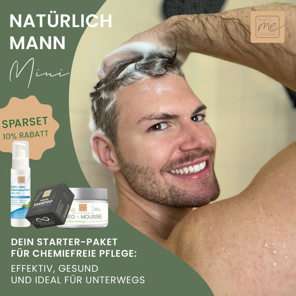 Ein lächelnder Mann wäscht sich die Haare, während er einen Duschkopf hält. Neben ihm stehen Thats me Organic® Deo-Mousse Lemongrass und Bio-Haarpflegeprodukte. Der deutsche Text beschreibt das perfekte Geschenk-Set für Männer.