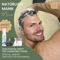Ein lächelnder Mann wäscht sich die Haare, während er einen Duschkopf hält. Neben ihm stehen Thats me Organic® Deo-Mousse Lemongrass und Bio-Haarpflegeprodukte. Der deutsche Text beschreibt das perfekte Geschenk-Set für Männer.